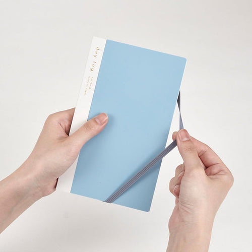 A person's hands holding a light blue notebook with a white spine and a gray elastic closure band, set against a plain off-white background.