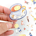 Hand holding sticker sheet with lemon tea and little human figures.