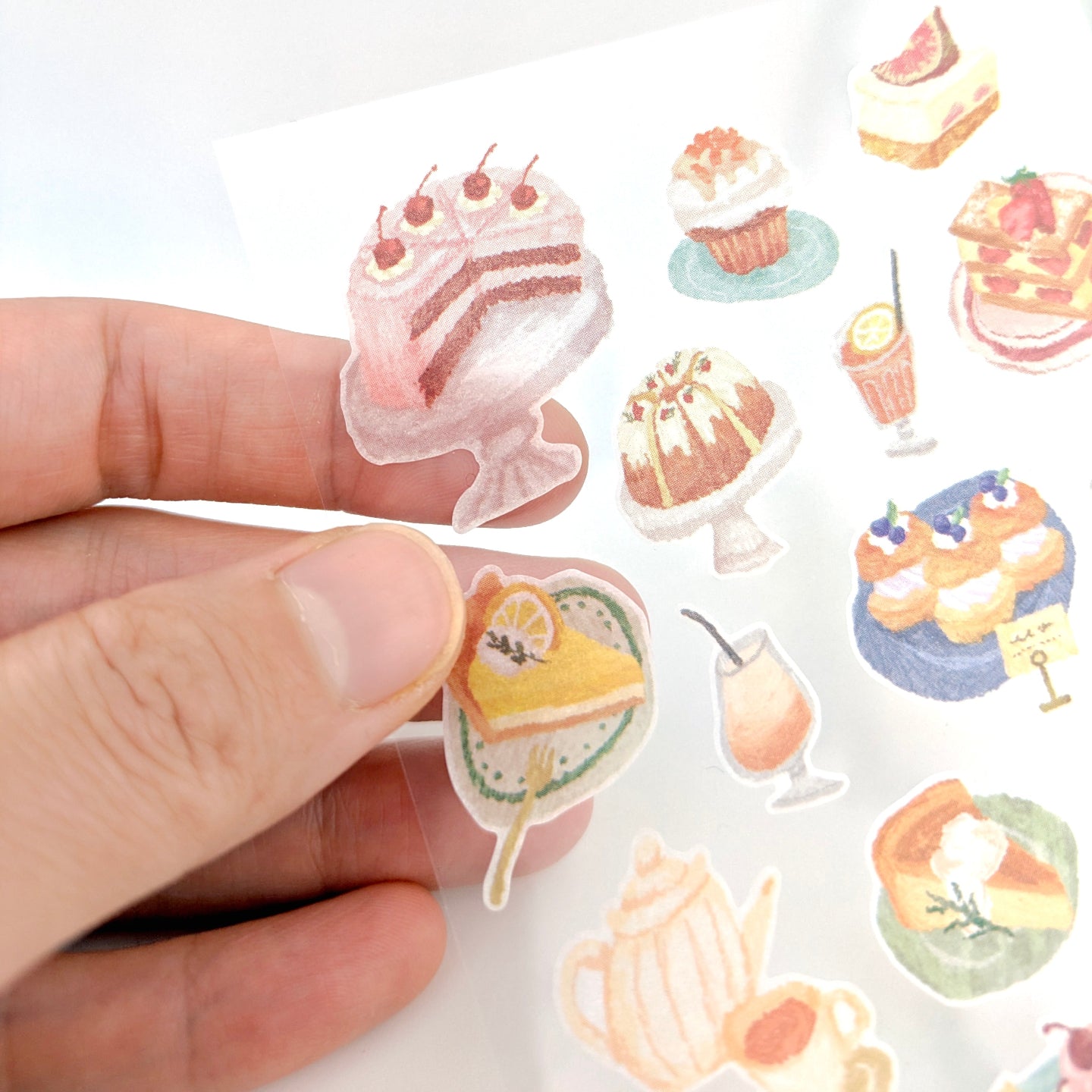 Hand holding a sheet of cake-themed stickers with various dessert and drinks.