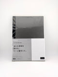 A slim rectangular notebook or journal with a dark charcoal gray cover and a light gray lower section featuring Japanese text and a white grid pattern, photographed on a clean white background.