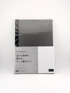 A slim rectangular notebook or journal with a dark charcoal gray cover and a light gray lower section featuring Japanese text and a white grid pattern, photographed on a clean white background.