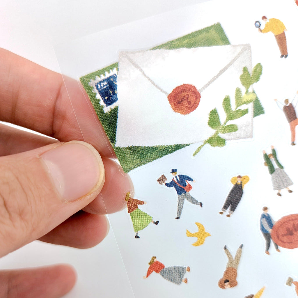 Hand holding a miniature envelope with illustrations of people and objects on a white background