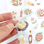 Hand holding a sticker of a dessert with more stickers of food items in the background.