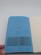 An open notebook or planner shown from the inside, with guide to use on a light blue page on the left, and a cream grid page on the right. A hand is holding the left edge of the book against a plain light background.