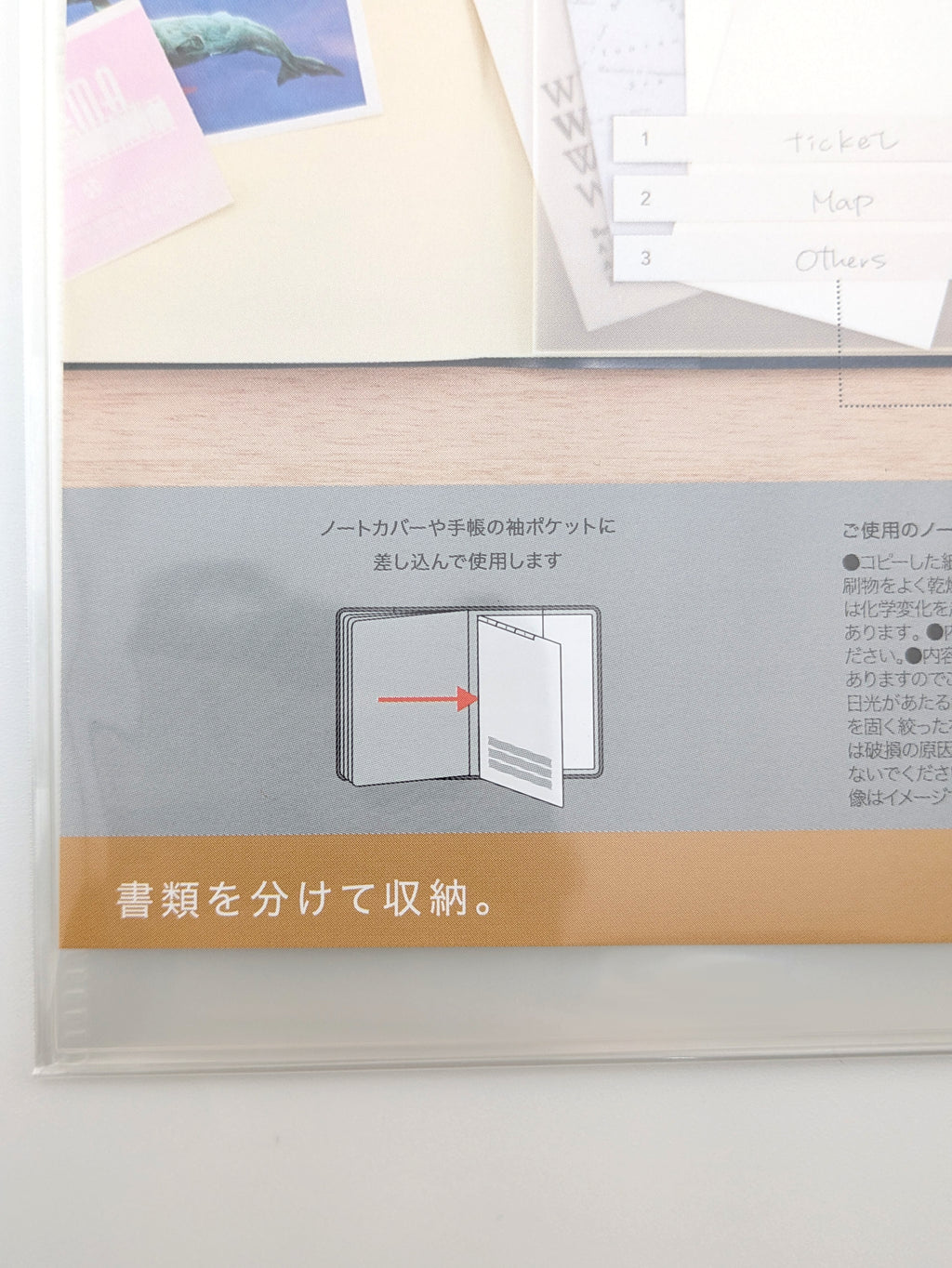A close-up of a clear plastic document organizer sleeve containing a light wood folder with a gray paper insert featuring Japanese instructions and a tan banner at the bottom.