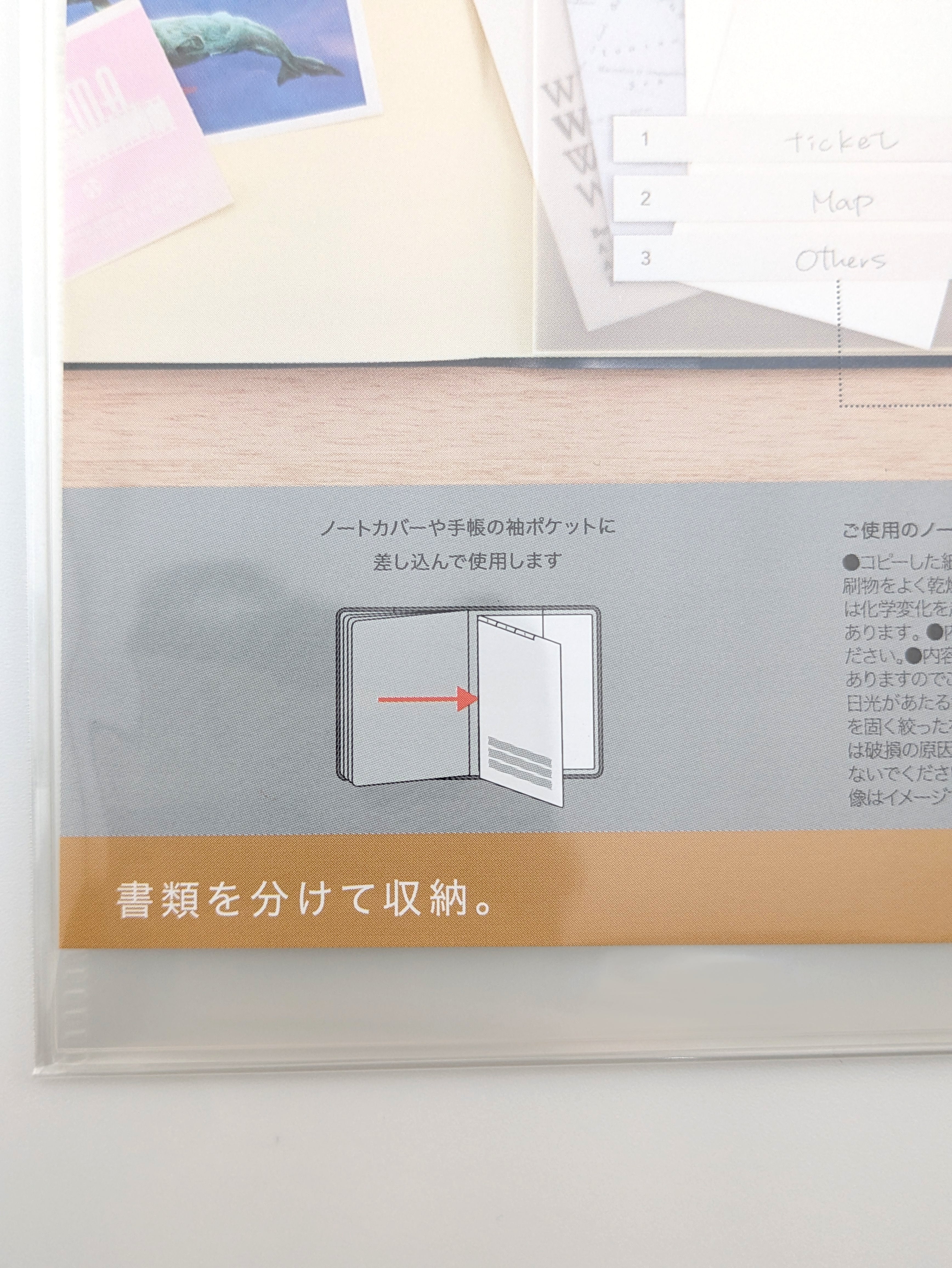 A close-up of a clear plastic document organizer sleeve containing a light wood folder with a gray paper insert featuring Japanese instructions and a tan banner at the bottom.