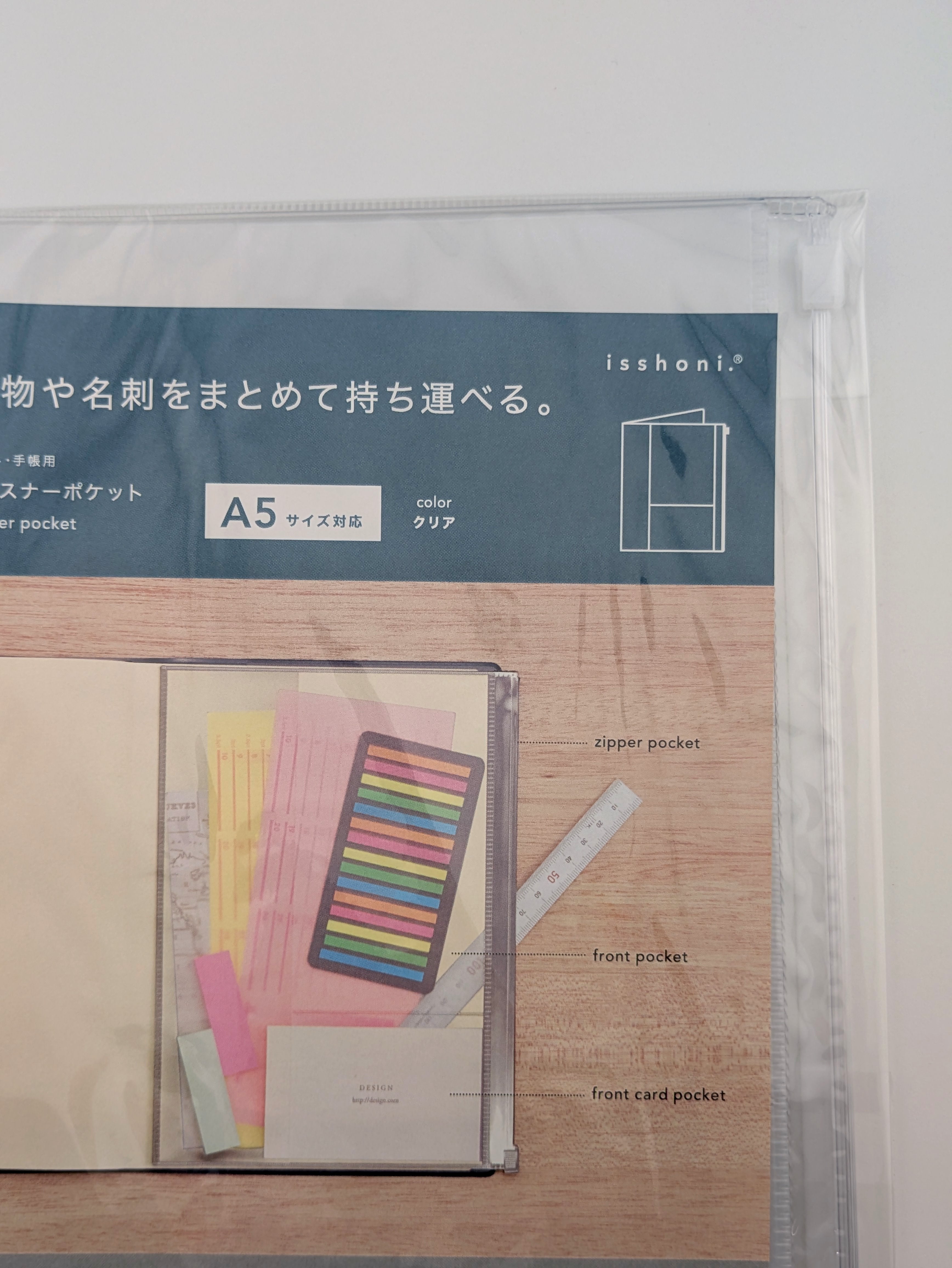 A packaged Isshoni A5 clear organizer pocket notebook shown in its retail sleeve, featuring a dark blue header with product details and a mockup of the notebook with pastel stationery and a ruler on a wood-grain background.